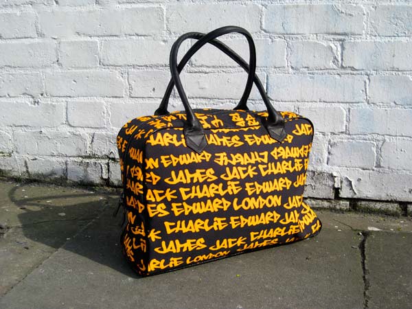 Black holdall with yellow graffiti text standing next to a white brick wall